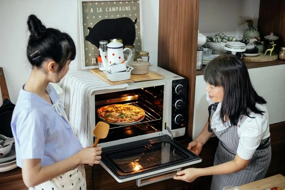 Focused Asian mother and daughter in aprons preparing to baking yummy homemade pizza in oven while cooking together in kitchen